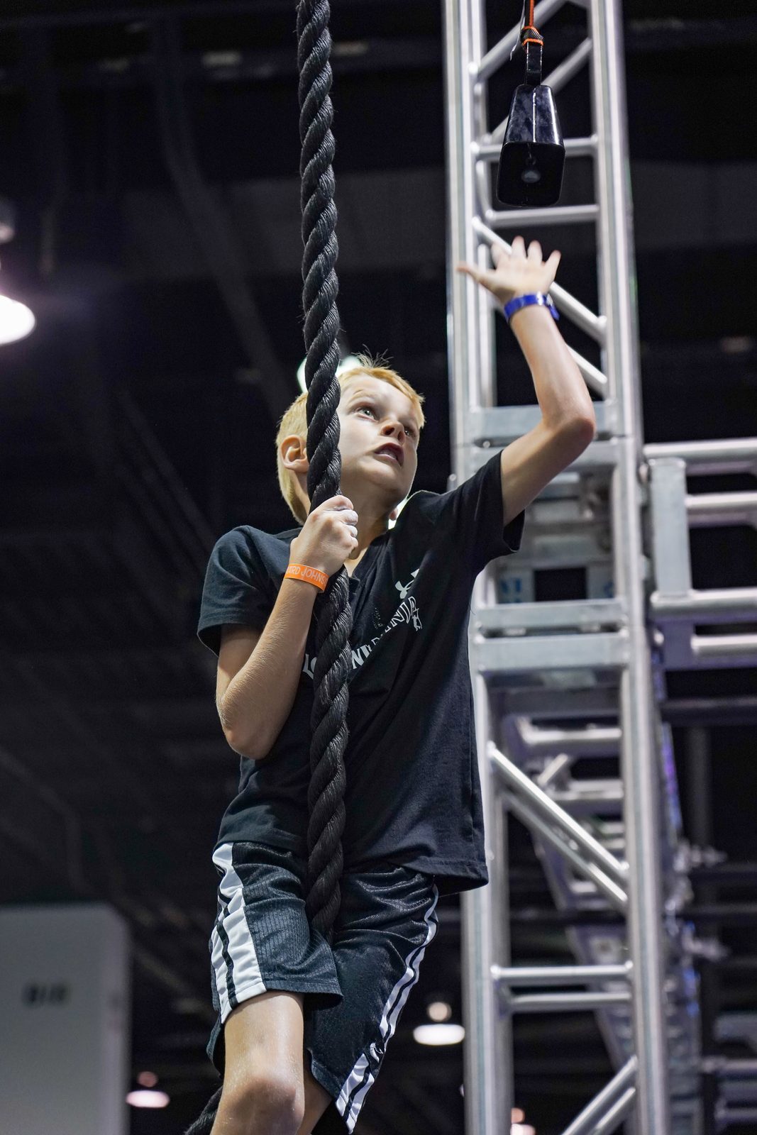 A boy climbing a rope high above the mats at a ninja league competition, focused, dark arena behind him