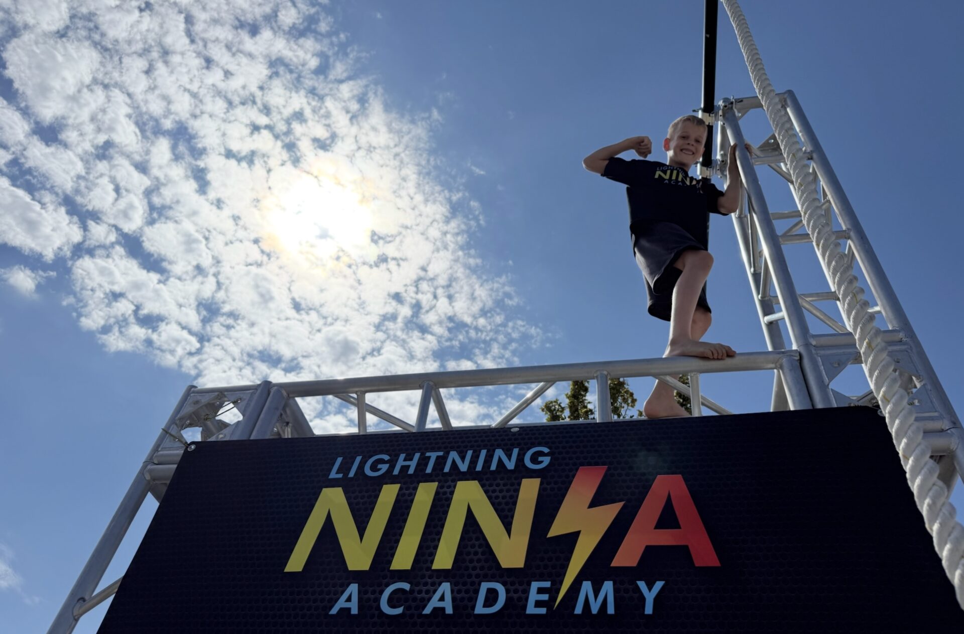 A kid flexing triumphantly on top of a truss rig under a blue sky, Lightning Ninja Academy banner below