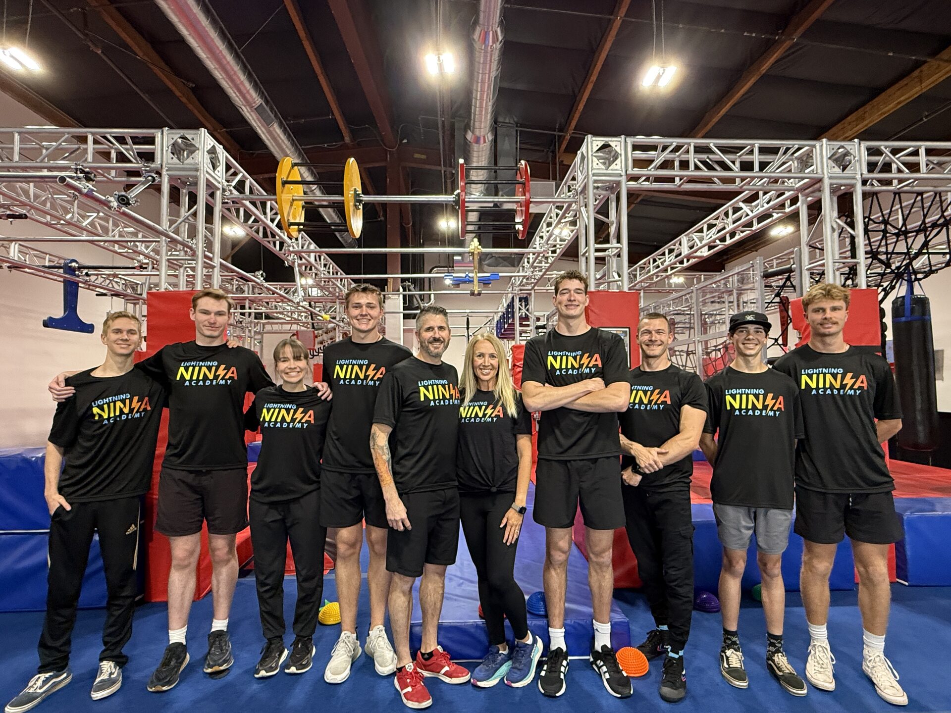 Lightning Ninja Academy's ten coaches smiling together on the gym floor, silver truss and obstacle rig behind them